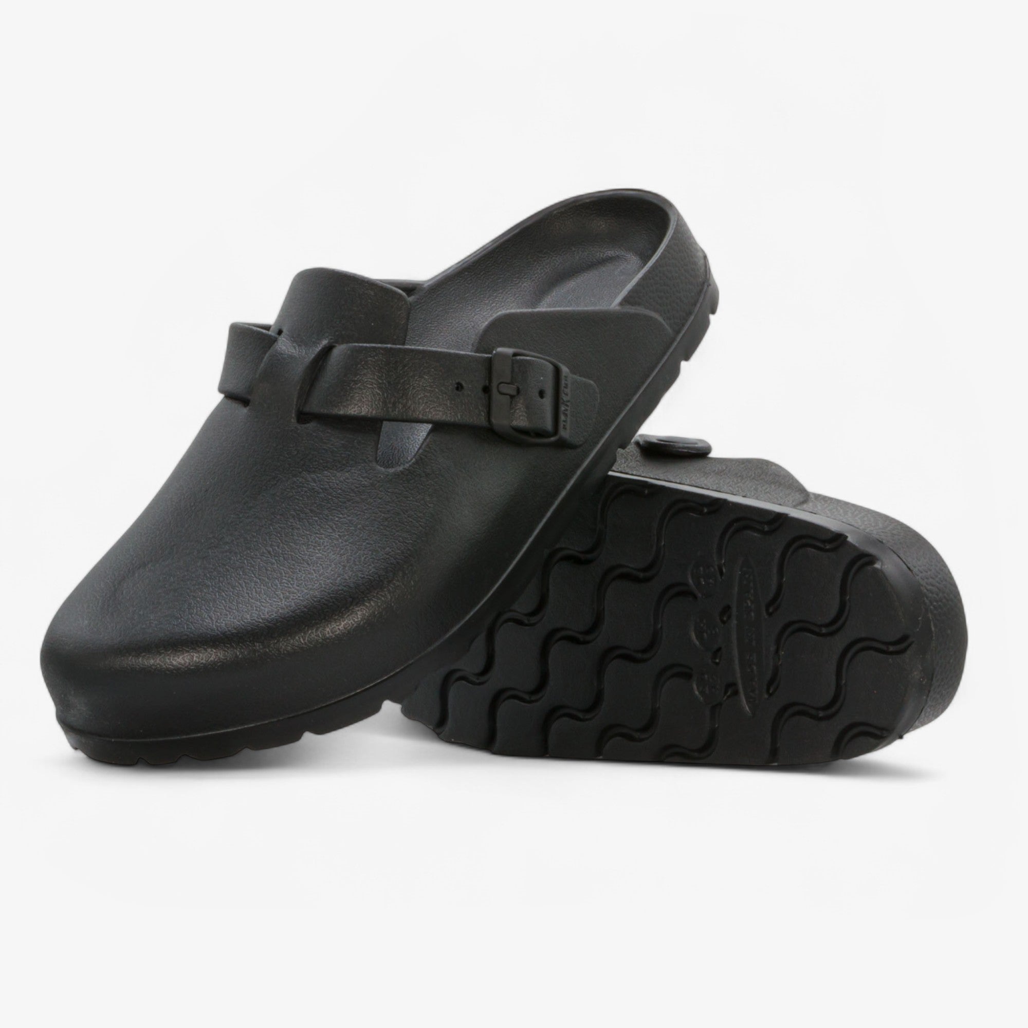A pair of black clog-style mules with a single strap and buckle, shown on a clean white background. One shoe is positioned upright to show the side profile, while the other lies on its side to display the textured outsole.